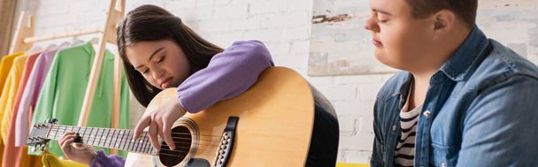 Έφηβος με σύνδρομο down παίζει ακουστική κιθάρα κοντά σε θολή φίλο στο σπίτι, banner - Φωτογραφία, εικόνα