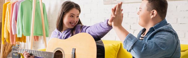 Cheerful teenagers with down syndrome and acoustic guitar giving high five at home, banner  - Photo, Image