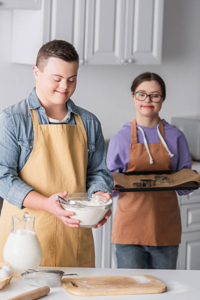 Lächelnder Teenager mit Down-Syndrom hält Mehl neben verschwommener Freundin mit Backblech in Küche  - Foto, Bild