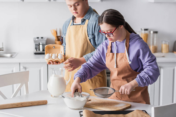 Girl with down syndrome cooking near friend holding eggs in kitchen  - Photo, Image