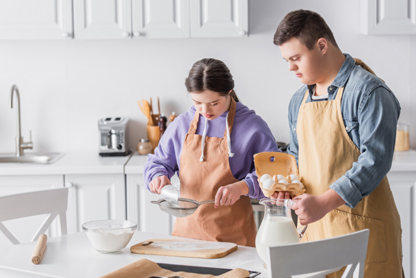 Tieners met dons syndroom koken in de buurt van melk en bloem in de keuken  - Foto, afbeelding
