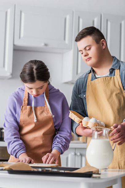 Girl with down syndrome cooking near friend with eggs and baking sheet in kitchen  - Photo, Image