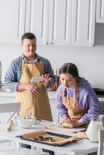 Teenager with down syndrome making dough near friend with smartphone in kitchen  - 写真・画像