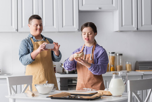 Teenie-Mädchen mit Down-Syndrom hält Teig neben Freundin mit Handy in Küche  - Foto, Bild