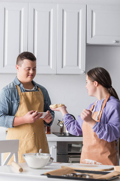 Positive teenager with down syndrome holding smartphone near friend with dough in kitchen  - Fotó, kép