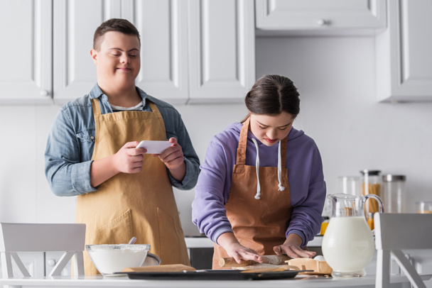 Adolescente con síndrome de Down haciendo masa cerca de los ingredientes y amigo sosteniendo teléfono inteligente en la cocina  - Foto, Imagen