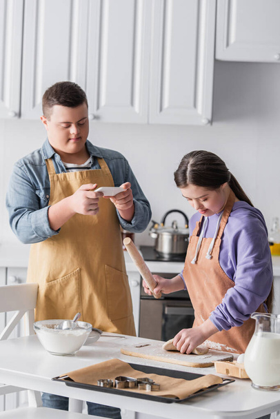 Adolescente con síndrome de Down cocinar masa cerca de amigo con teléfono inteligente en casa  - Foto, imagen