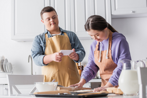 Nastoletni chłopiec z zespołem Downa trzymając smartfona podczas zamazany przyjaciel toczenia ciasta w kuchni  - Zdjęcie, obraz