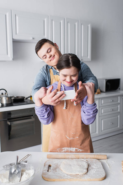 Teenager with down syndrome holding cellphone near friend in apron and dough in kitchen  - 写真・画像