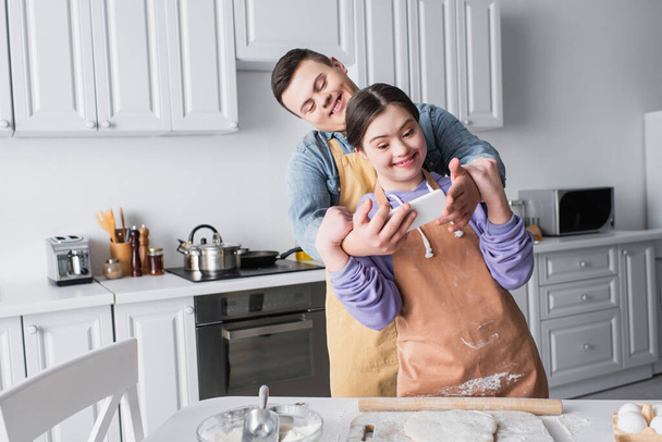 Alegre pareja con síndrome de Down tomando selfie en el teléfono inteligente cerca de la masa en la cocina  - Foto, Imagen