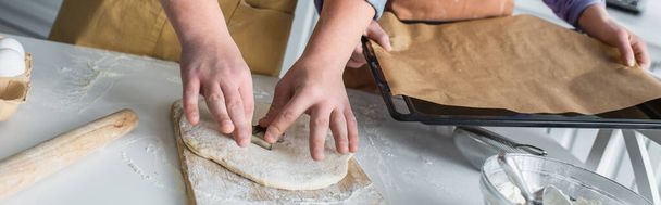 Cropped view of teenager making cookie near friend with baking sheet at home, banner  - Foto, imagen