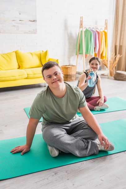 Sonriente adolescente con síndrome de Down mirando a la cámara en la alfombra de fitness en casa  - Foto, imagen