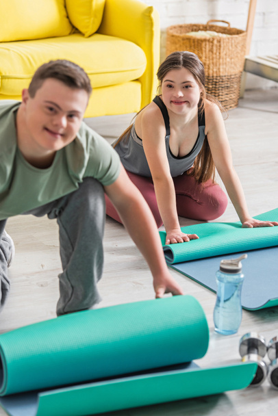 Teenager with down syndrome sitting near fitness mat and blurred friend at home  - Fotoğraf, Görsel