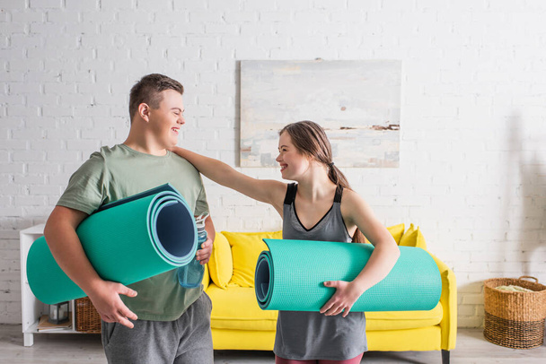 Positive teenager with down syndrome holding fitness mat near boyfriend with sports bottle at home  - Zdjęcie, obraz
