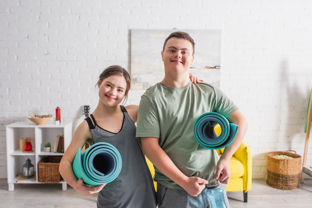 Θετικό ζευγάρι εφήβων με σύνδρομο down κρατώντας πατάκια γυμναστικής στο σπίτι  - Φωτογραφία, εικόνα
