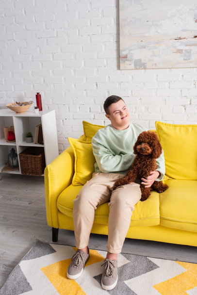 Cheerful boy with down syndrome petting poodle on couch at home  - Zdjęcie, obraz