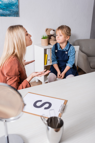 Terapeuta del habla trabajando con un niño cerca de espejo borroso y carta en la sala de consulta  - Foto, Imagen
