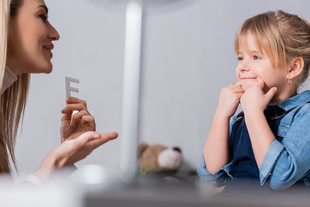 Enfant souriant touchant la bouche près de l'orthophoniste avec lettre dans la salle de consultation  - Photo, image