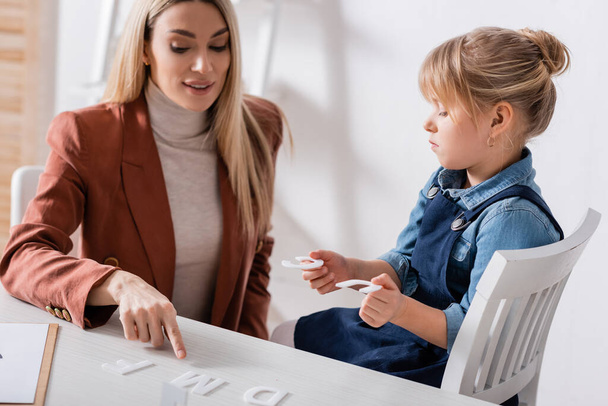 Speech therapist pointing at letter on table near kid in consulting room  - Foto, imagen