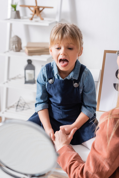 Child talking near blurred mirror and speech therapist with clipboard in consulting room  - 写真・画像
