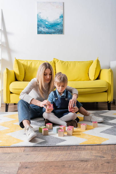 Kid holding wooden blocks with letters near speech therapist on carpet in consulting room  - Φωτογραφία, εικόνα