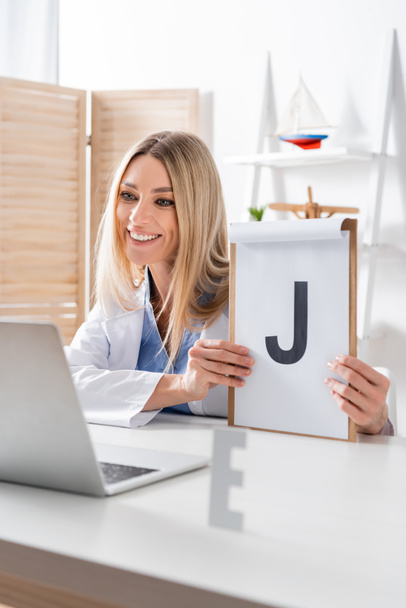 Happy logopedist holding clipboard during video call on laptop in consulting room  - Фото, изображение