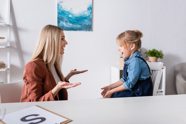 Side view of cheerful speech therapist talking with girl in consulting room  - Photo, Image