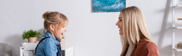Vista laterale del bambino che parla vicino al logopedista sorridente nella sala di consultazione, banner  - Foto, immagini