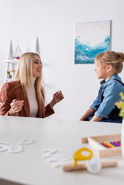 Logopedist parler avec fille souriante près de lettres floues et entraîneur de muscle respiratoire dans la salle de consultation  - Photo, image
