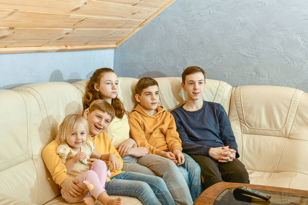 Cinco niños se sientan en un sofá de cuero y ven la televisión. - Foto, Imagen
