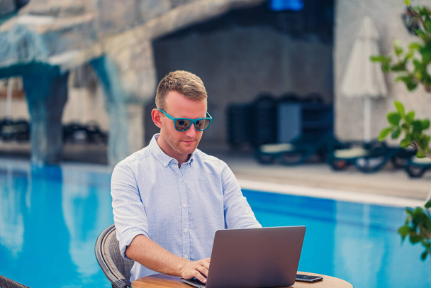 Młody biznesmen w okularach przeciwsłonecznych pracuje przy laptopie przy stole przy basenie. Praca zdalna. Wolny strzelec - Zdjęcie, obraz