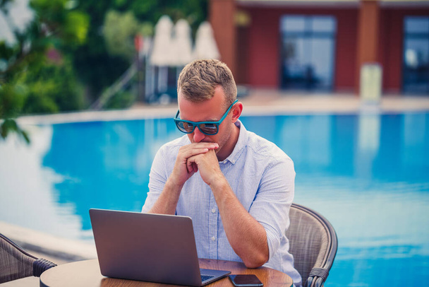 Der junge männliche Geschäftsmann mit Sonnenbrille sitzt an einem Laptop an einem Tisch in der Nähe des Pools. Fernbedienung. Freiberufler - Foto, Bild