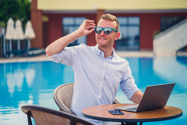Joven hombre de negocios con gafas de sol trabaja en un portátil sentado en una mesa cerca de la piscina. Trabajo remoto. Autónomo - Foto, imagen