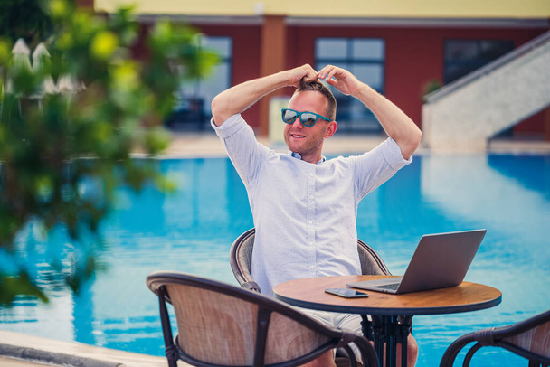 Udany przystojny biznesmen w okularach przeciwsłonecznych pracuje w laptopie, siedząc przy basenie. Praca zdalna. Wolny strzelec - Zdjęcie, obraz