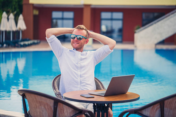 El exitoso hombre de negocios guapo con gafas de sol trabaja en un portátil sentado cerca de la piscina. Trabajo remoto. Autónomo - Foto, Imagen