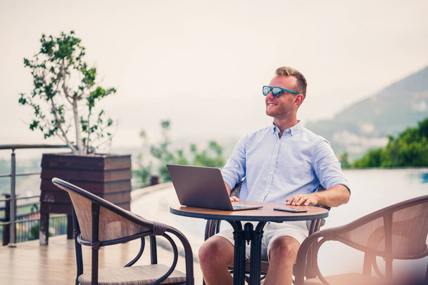 El exitoso hombre de negocios guapo con gafas de sol trabaja en un portátil sentado cerca de la piscina. Trabajo remoto. Autónomo - Foto, imagen