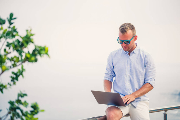 Fiatal, sikeres férfi üzletember, aki laptoppal dolgozik a tengerparti nyaraláson. Napszemüveget, inget és fehér nadrágot visel. Dolgozz az irodán kívül, szabadúszó. - Fotó, kép