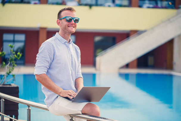 Jeune homme d'affaires avec ordinateur portable assis près de la piscine. Lieu de travail ouvert. Concept de pigiste. Achats en ligne. Travaux de vacances - Photo, image