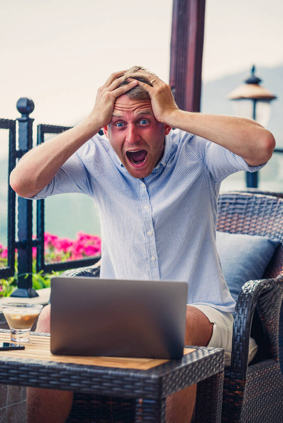 Un hombre emocional se sienta en la computadora de vacaciones y trabaja. Freelancer trabaja de vacaciones. Foto de alta calidad - Foto, Imagen
