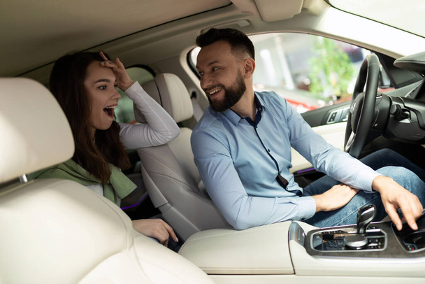 um homem e uma mulher em uma concessionária de carros estão sentados dentro do carro, sendo surpreendidos por novas tecnologias e promoções - Foto, Imagem