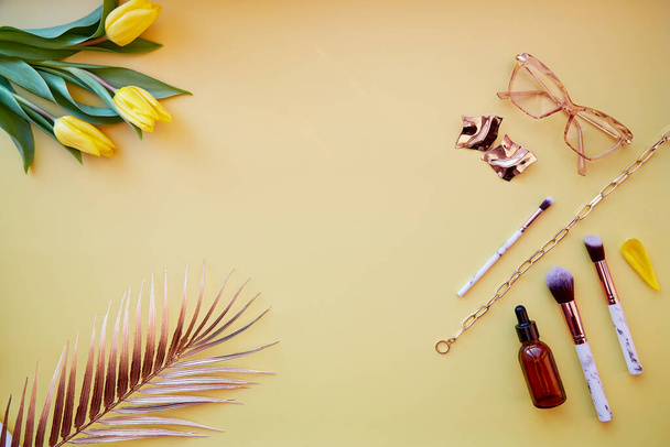 Cosméticos de moda, accesorios, pinceles de maquillaje sobre fondo amarillo pastel. Hojas de palma, tulipanes amarillos, gafas, gotero de suero. Copiar espacio - Foto, imagen