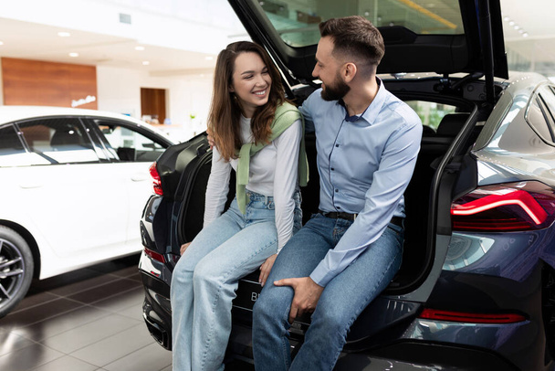 szczęśliwie młoda para siedzi w bagażniku samochodu zwisając nogami w salonie samochodowym - Zdjęcie, obraz