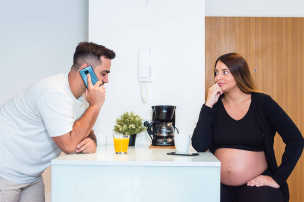 mladý heterosexuální těhotný pár doma spolu ráno komunikují, pijí kávu a pomerančový džus, manžel mluví po telefonu a žena ho pozoruje. - Fotografie, Obrázek