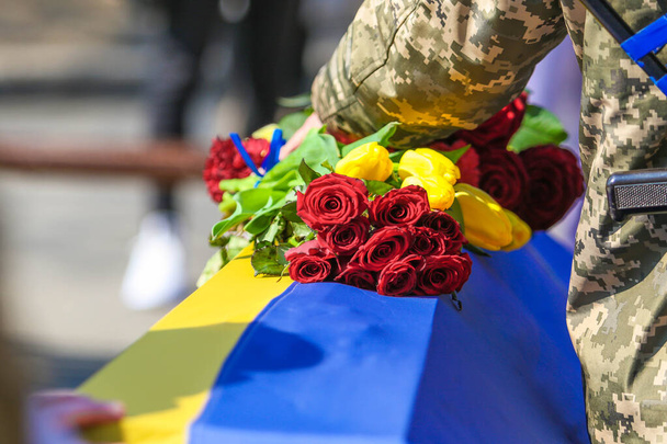 POLTAVA, UKRAINE - 24. MÄRZ 2022: Ein Soldat hält Blumen auf einem mit der ukrainischen Flagge bedeckten Grab während der Beisetzungszeremonie für gefallene Soldaten der ukrainischen Streitkräfte - Foto, Bild