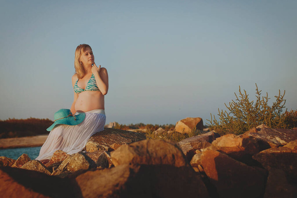 Krásná mladá elegantní těhotná blondýna v modrém klobouku kráčí po pláži, na pobřeží při západu slunce. Azovské moře. Mateřství. Mateřství. - Fotografie, Obrázek