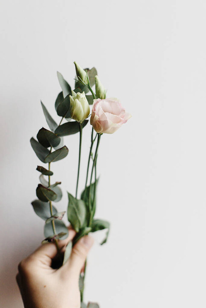 Hand holding spring modern bouquet on white background close up. Beautiful fresh flowers eustoma and eucalyptus. Stylish minimal floral arrangement, moody image - Fotografie, Obrázek