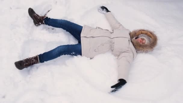 Una mujer con una chaqueta blanca yace en la nieve, levanta las manos hacia arriba y hacia abajo, como si agitara sus alas y hiciera un ángel - Metraje, vídeo