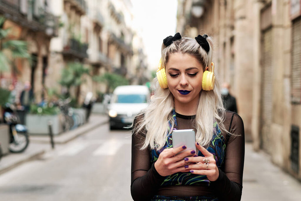 Młoda kobieta alternatywna korzystająca z telefonu komórkowego ze słuchawkami podczas spaceru ulicą. - Zdjęcie, obraz
