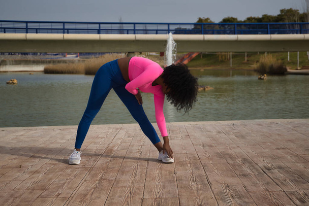 Афро-американка с афро-волосами и спортивной одеждой, с флуоресцентной розовой футболкой и леггинсами, растянувшейся у озера на открытом воздухе. Концепция, спорт, улица, город. - Фото, изображение