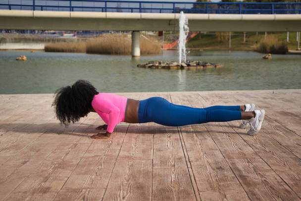 African-American woman with afro hair and sportswear, with fluorescent pink t-shirt and leggings, doing push-ups next to an outdoor lake. Fitness concept, sport, street, urban. - Photo, Image
